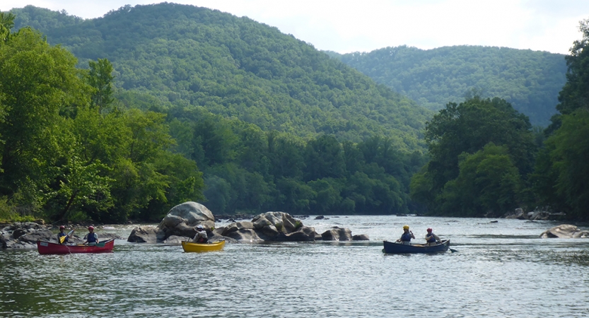 Three canoes, each carrying two people, float on a river amid rock formations. In the background, mountains are covered in thick green trees.